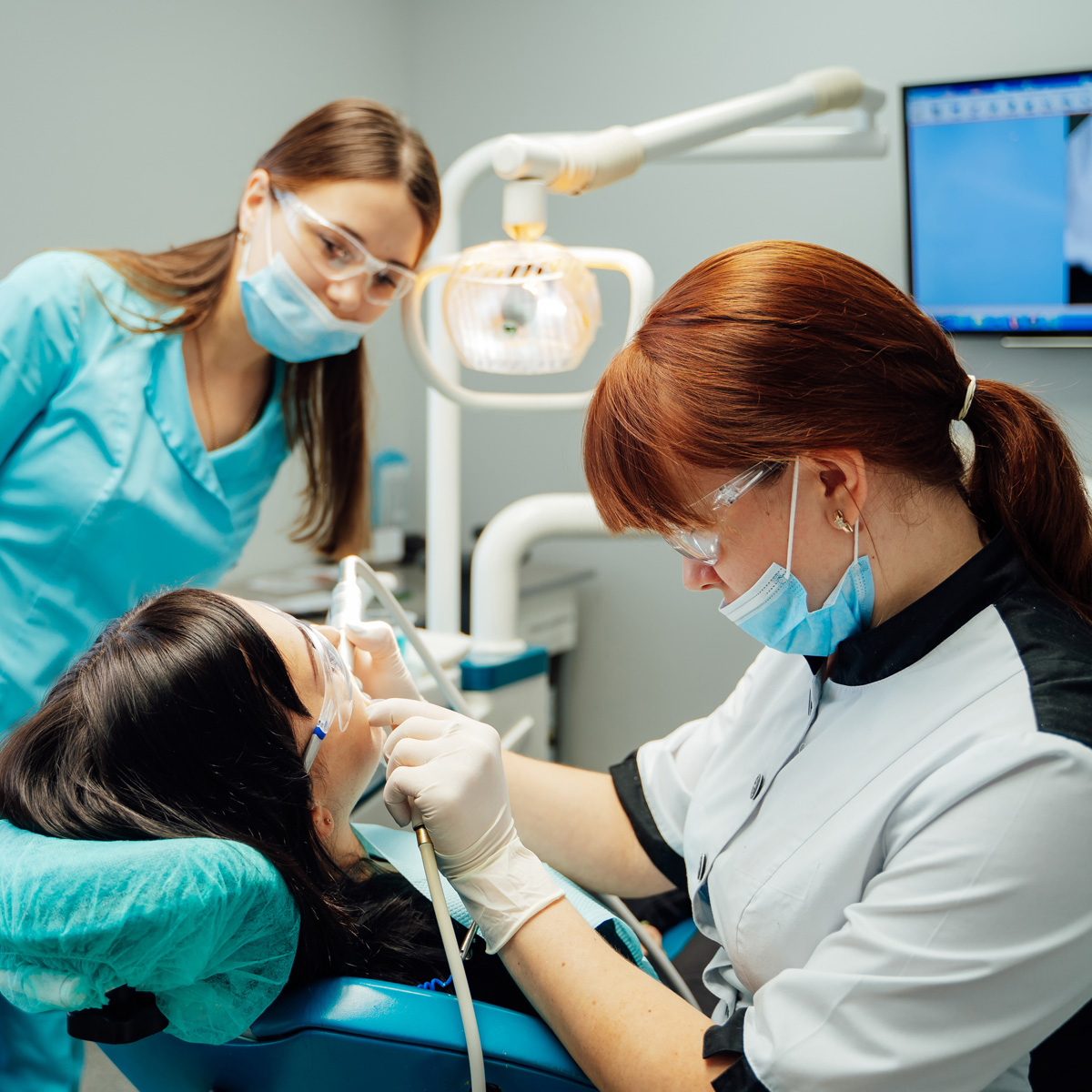 Two dental professionals assist a patient during an oral treatment in a modern clinic setting.