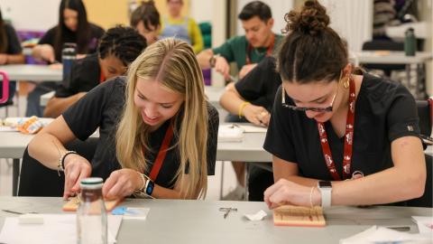 A pair of students are seen sitting and practicing stitching skills at a desk.