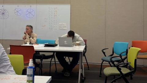Adults sit at a desk. There's a whiteboard with math on it behind them.