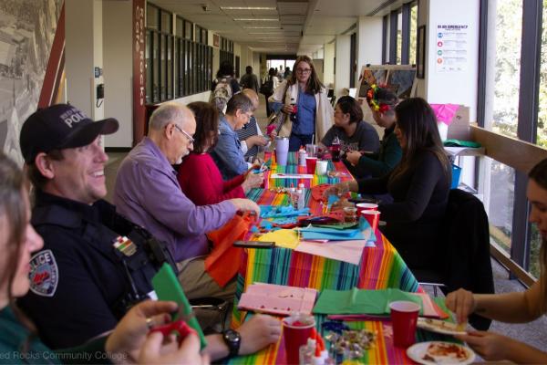 People sit around colorful tables, cutting paper and eating salsa, in a hallway at RRCC.