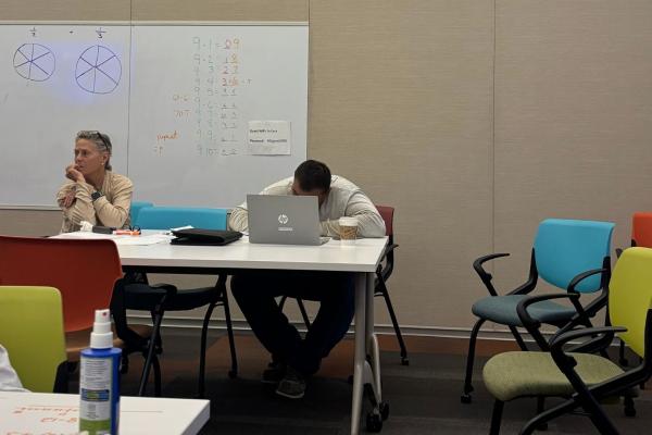 Adults sit at a desk. There's a whiteboard with math on it behind them.