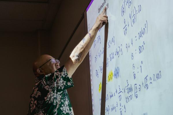 A man in glasses and a Hawaiian shirt points at a white board with math problems on it.