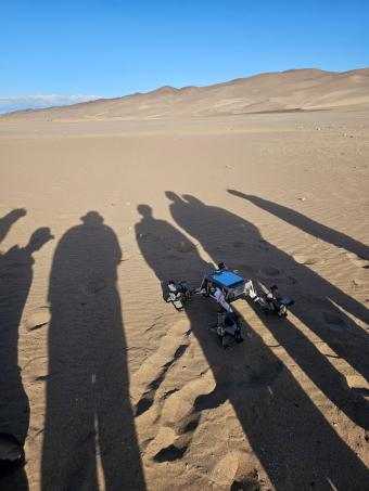 a robot sits on sand and the shadow of several people is seen over it