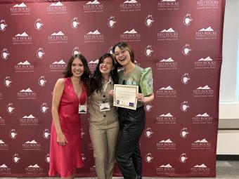 Three women pose in front of a backdrop with the RRCC logo and hold an award.