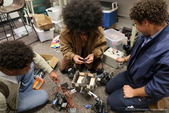 Three students sit on the floor working on a robot that sits in the middle
