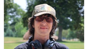 Profile photo of Sam who is outdoors, smiling and wearing a hat and glasses