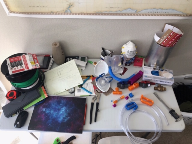 Home setup for 3D printing workspace. Various different pieces of equipment on top of table, including reem of green plastic rope for 3D printing, dust masks, plastic tubes, hand tools, papers, and binders. 